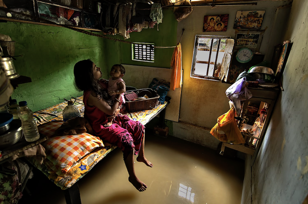 Mother and baby seated on a bed, above a flooded floor in their small home, possessions raised above the water.