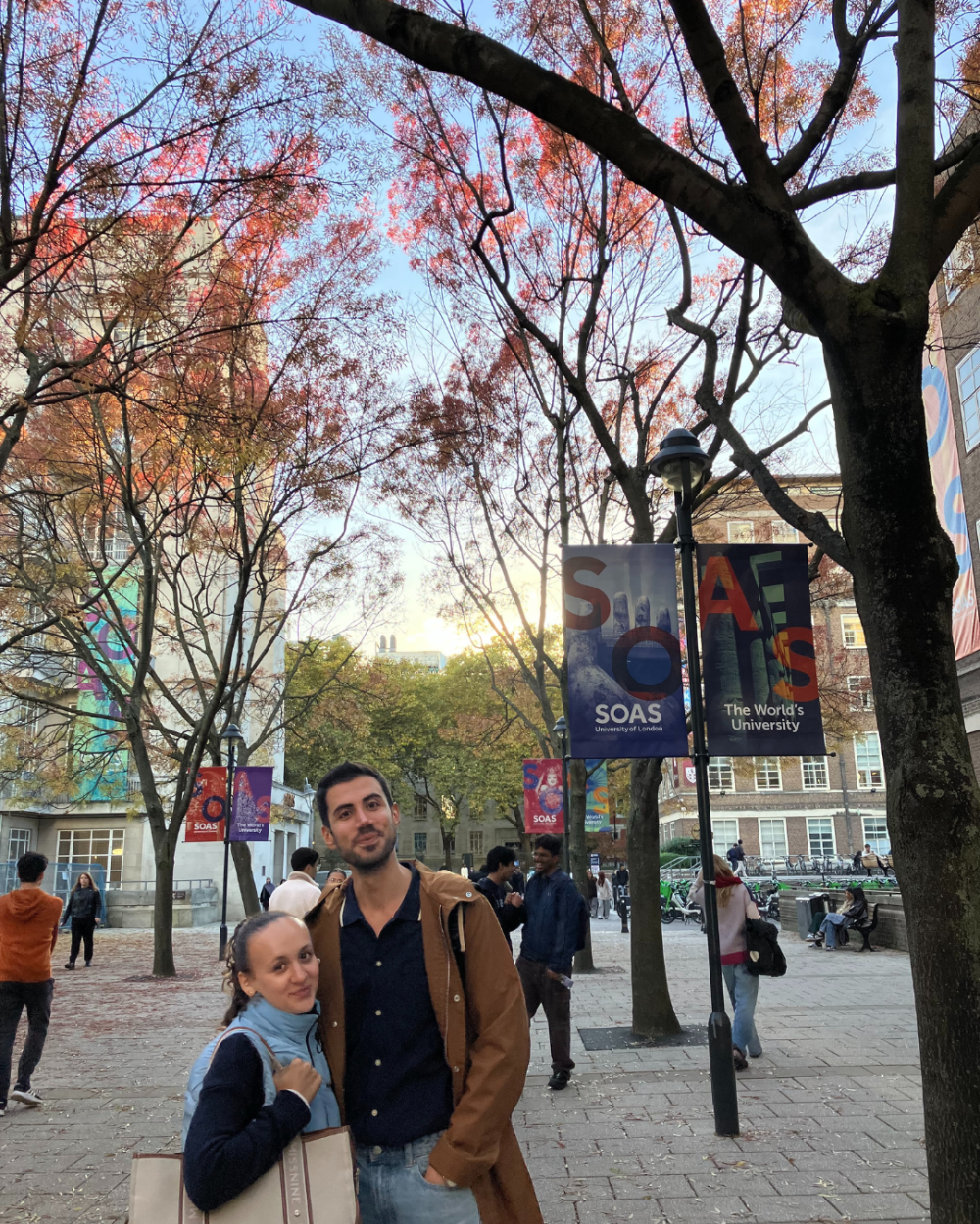 Two students smile at the camer underneath the autumnal trees and colourful SOAS signs